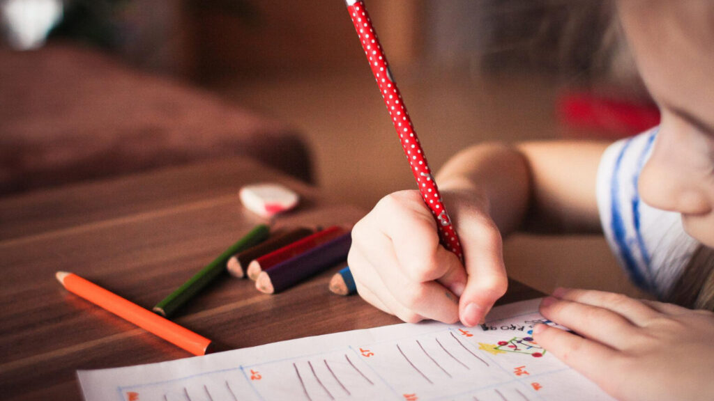 Lernen (Symbolbild): Junger Schüler sitzt mit großem Bleistift in dern kleinen Hand vor einem Arbeitsblatt, das er konzentriert bearbeitet