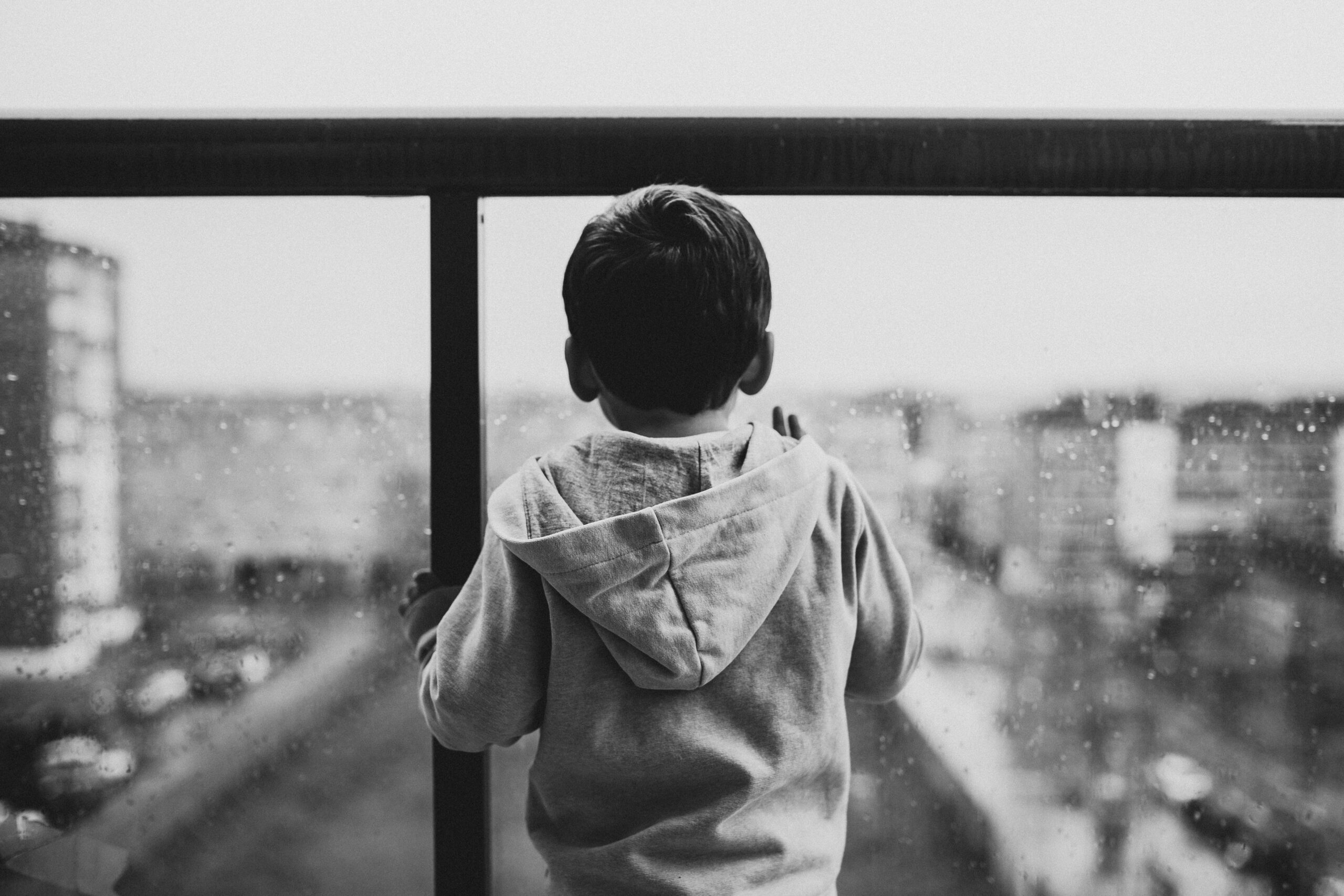 Rückansicht eines traurigen Kindes, das durch eine gläserne Balkonbrüstung auf eine Stadt guckt. Foto von Juan Pablo Serrano Arenas: https://www.pexels.com/de-de/foto/ruckansicht-eines-traurigen-kindes-das-auf-ein-glasfenster-wartet-827993/