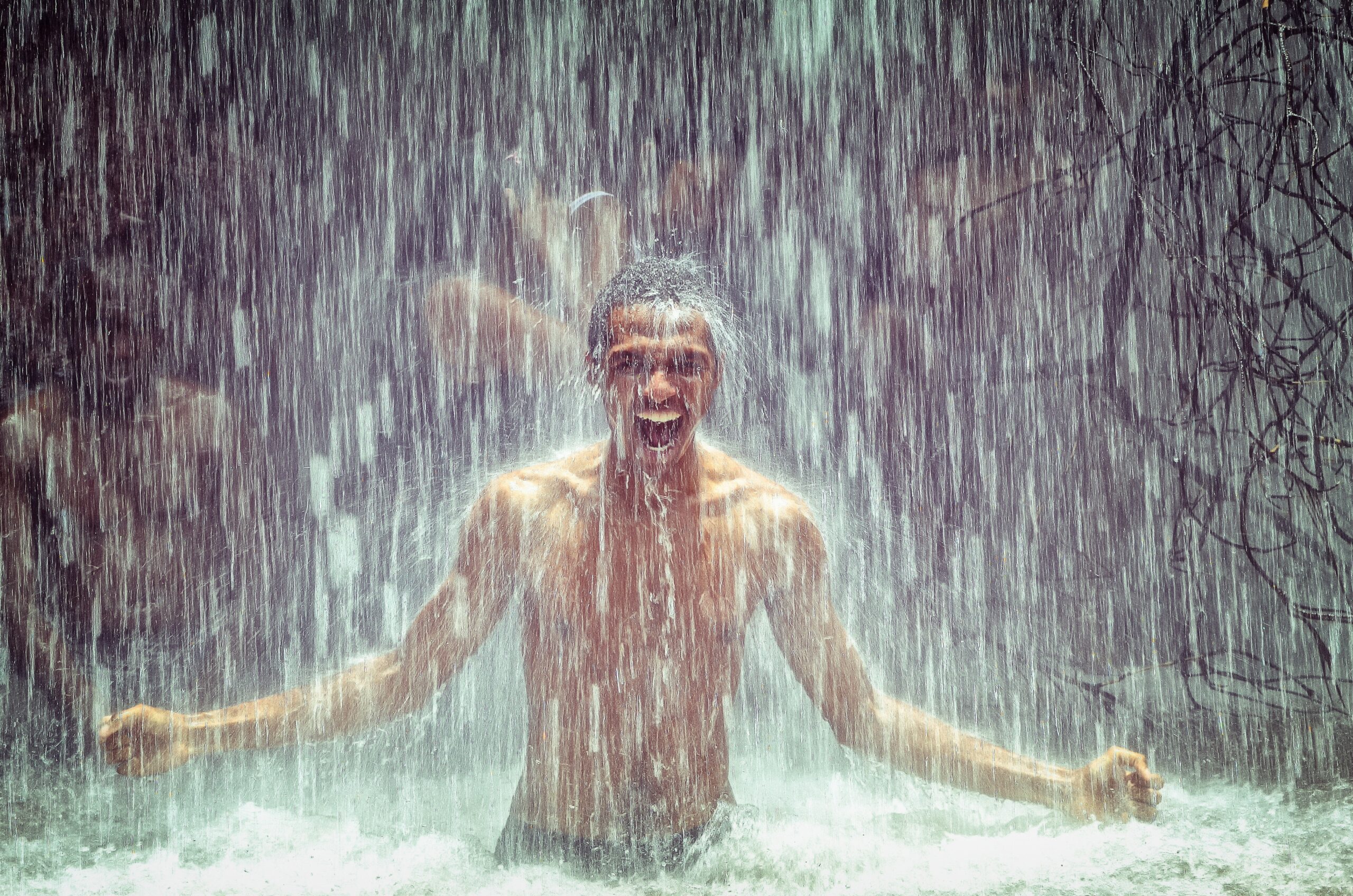 Männliche PoC duscht freudestrahlend im Wasser eines Wasserfalls. In Hintergrund sind verschwommen weitere Personen zu erkennen.