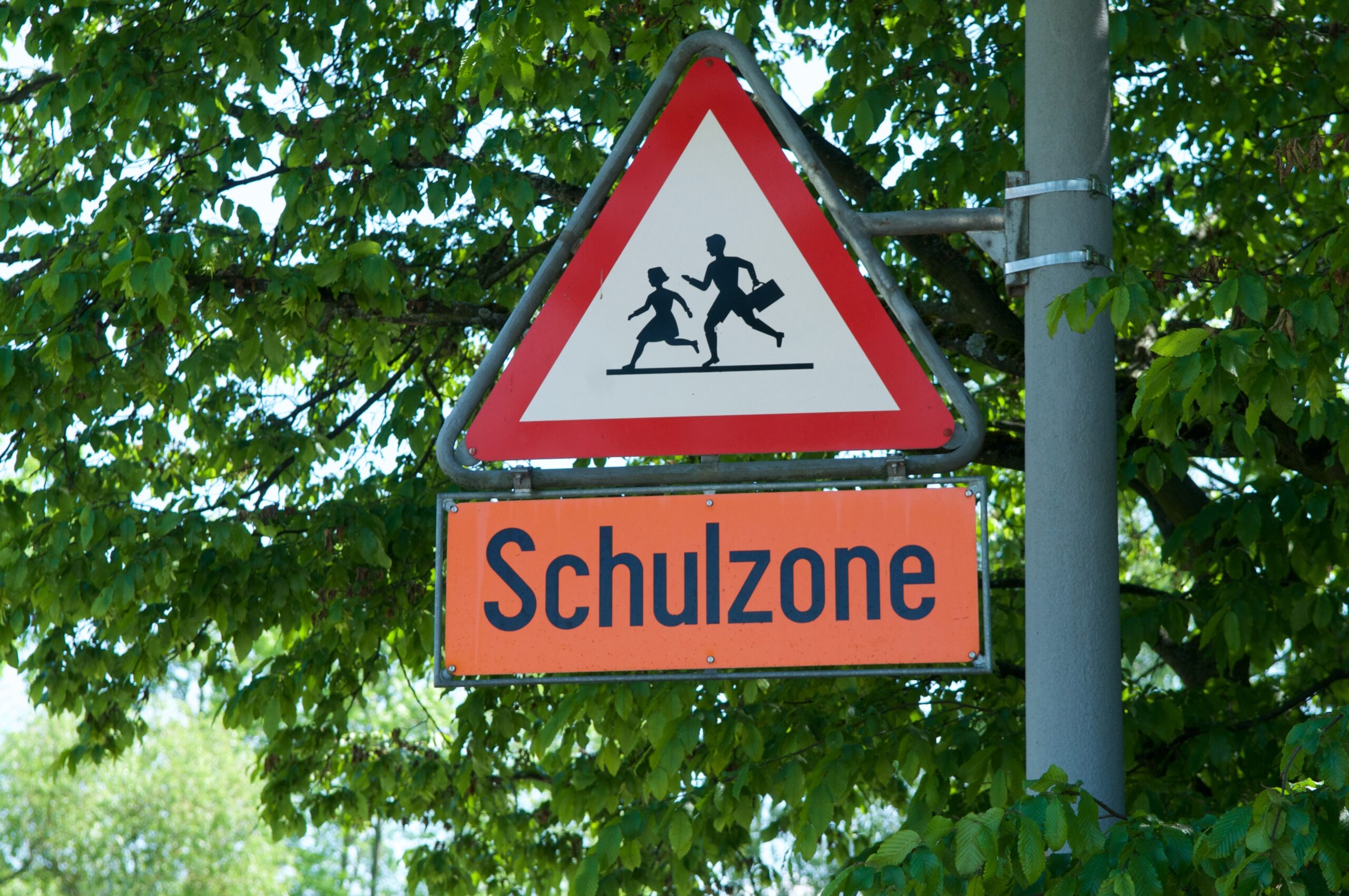 Das Verkehrszeichen 136 Kinder hängt an einer Laterne vor einer Baumkrone, darunter ein oranges Hinweisschild "Schulzone"